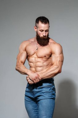 Atletik yakışıklı sakallı, kaslı, çıplak, mavi kot pantolonlu, gri arka planda yalnız bir adam..