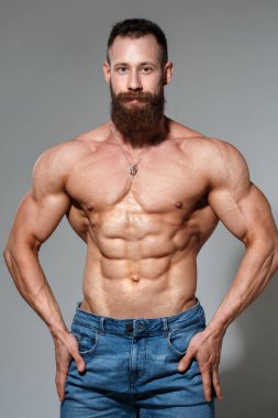 Atletik yakışıklı sakallı, kaslı, çıplak, mavi kot pantolonlu, gri arka planda yalnız bir adam..