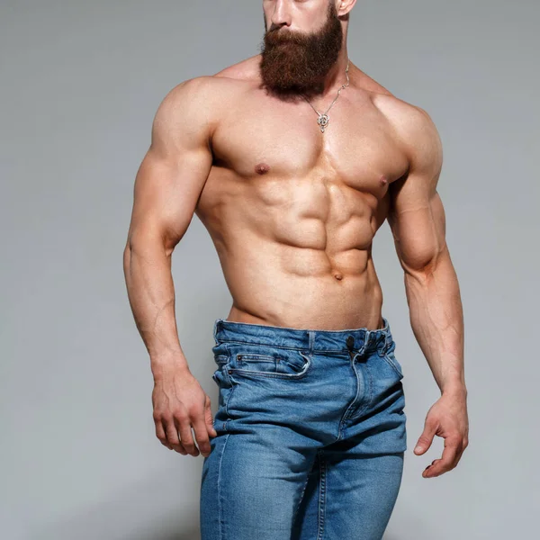 Atletik yakışıklı sakallı, kaslı, çıplak, mavi kot pantolonlu, gri arka planda yalnız bir adam..