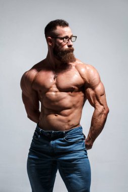 Gri bir arka planda çıplak gövdesi olan sakallı ve gözlüklü kaslı genç bir adam..