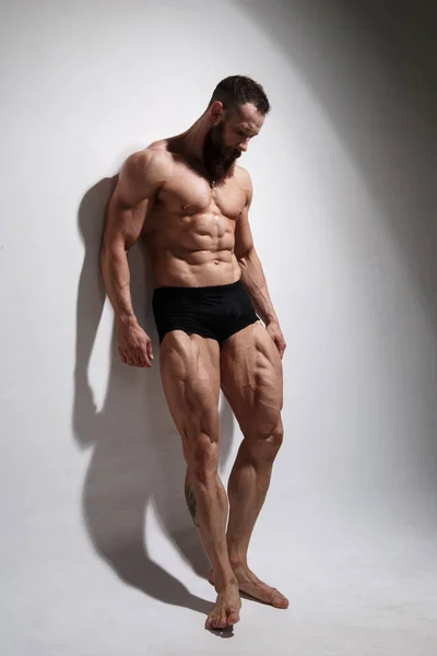 Athletic bearded man shows muscles standing in full growth on a light background.