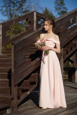 Genç bir kız elinde buketi olan zarif pembe elbiseli gelin yaz günü kırsal bir terasta tam gelişim içinde duruyor..