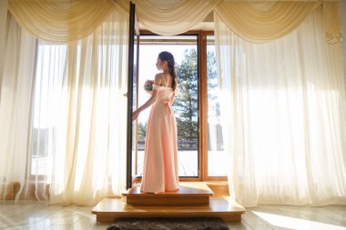 Yumuşak pembe elbiseli genç bir gelin elinde bir buketle güneşli bir günde terasın kapısında dikiliyor..