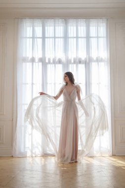 İnce bir gelinin zarif silueti, akıcı bir elbiseyle pencerenin arka planında..