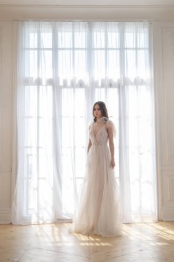 İnce bir gelinin zarif silueti, akıcı bir elbiseyle pencerenin arka planında..