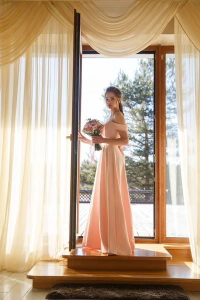 在一个阳光灿烂的日子里 一位年轻的新娘站在阳台的门口 她身穿柔软的粉色连衣裙 手里拿着一束鲜花 — 图库照片