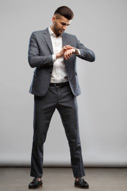 Zarif takım elbiseli genç şık bir adam gri bir arka planda tamamen büyümüş olarak saatine bakıyor..