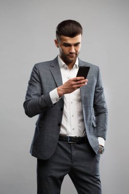 Zarif takım elbiseli şık genç adam elinde gri bir arka planda izole edilmiş bir akıllı telefon tutuyor..