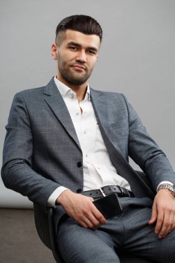 Gri bir arka planda tek başına oturan takım elbiseli karizmatik Asyalı adam..