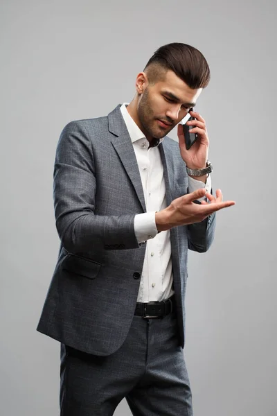 Stylish young man in an elegant suit talking on the phone gesturing with hands isolated on a gray background.
