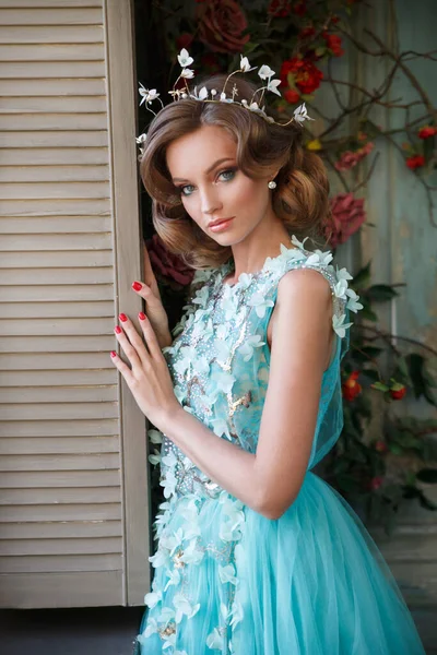 Beautiful girl in an airy turquoise dress on the terrace near a blooming rose bush.