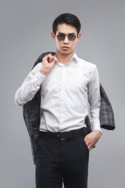 Güneş gözlüklü genç Asyalı bir adam elinde gri arka planda izole edilmiş bir ceket tutuyor..