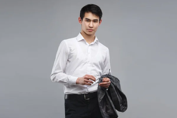 A young Asian man in a white shirt is holding a jacket isolated on a gray background.