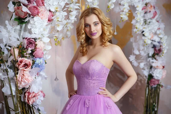 Güzel kız, güzel leylak elbiseli ve makyajlı sarışın çiçeklerin dekoratif kemerinin yanında..
