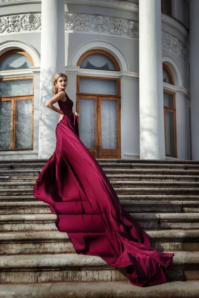 美しいです細い女の子で暗い赤い絹の流れるドレスに長い列車で宮殿の階段 — ストック写真