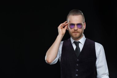 Sakallı yakışıklı adam güneş gözlüklerini düzeltiyor. Siyah arka planda izole edilmiş..