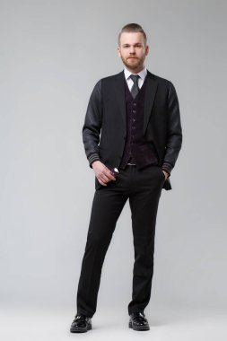 Üç parçalı takım elbiseli şık bir adam gri bir arka planda tam gelişim içinde duruyor..