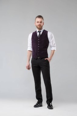 Yelek ve kravat takmış zarif bir adam gri bir arka planda duruyor..