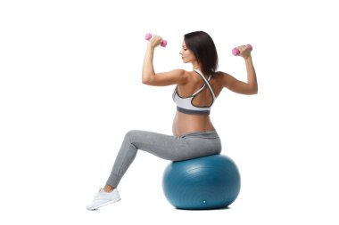 İnce atletik hamile kız, beyaz arka planda izole bir şekilde top görüşünde oturan dambıllarla spor yapıyor..