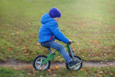 Mavi ceketli küçük bir çocuk sonbahar parkında koşan bir çantaya biniyor..