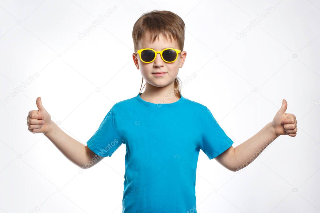 A little boy in blue clothes and sunglasses shows on both hands a sign a thumb raised up, isolated.