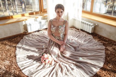 Güneşli bir günde, otel odasında panoramik pencereleri olan bir odada uzun eteği olan güzel bir gelin yerde, bir otel odasında oturuyor..