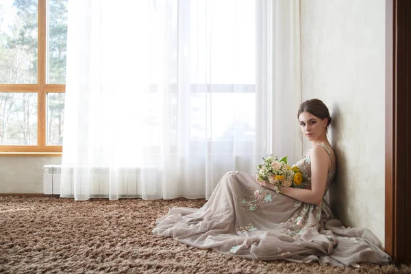 一个穿着雅致衣服的漂亮姑娘新娘坐在一间明亮的房间里的地毯上 房间有很大的窗户 — 图库照片