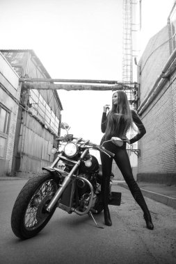 Endüstriyel bir caddede motosikletin yanında güzel, narin bir kızın siyah beyaz fotoğrafı..