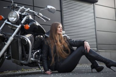 İnce, güzel, uzun saçlı, motosikletli bir kız, endüstriyel bir şehir binasının arka planında motosikletin yanında oturuyor..