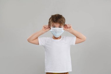 Tıbbi maskeli küçük bir çocuk iki elini de kafasına kaldırdı ve gri arka planda izole edilmiş boynuzlar çizdi..