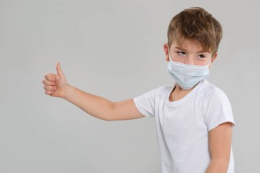 Yüzünde mutsuz bir dejenerasyon olan maske takan küçük bir çocuk gri arka planda izole edilmiş bir baş parmak gösteriyor..