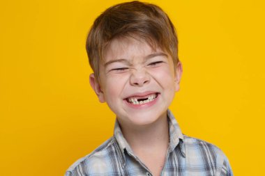 Little cute boy in a plaid shirt emotionally grins showing teeth isolated on a yellow background.