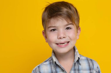 Little cute boy in a plaid shirt emotionally grins showing teeth isolated on a yellow background.