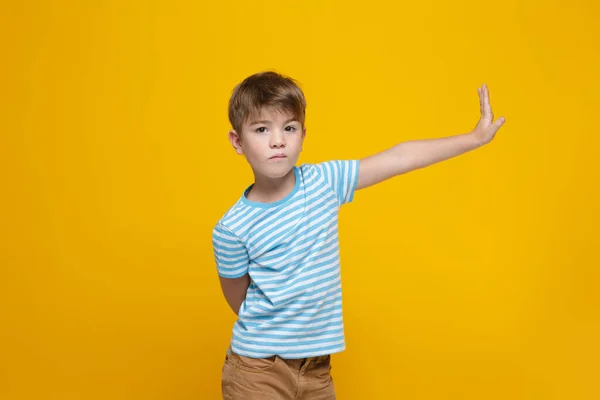 A little cute boy in a blue and white striped T-shirt stretches out his right hand with the palm raised in a prohibitory gesture, isolated on a yellow background.