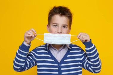 Mavi çizgili süveter giyen küçük şirin çocuk koruyucu bir maske takıyor, sarı arka planda izole edilmiş bir şekilde..