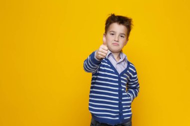 Çizgili süveter giyen küçük komik bir çocuk, parlak sarı bir arka planda izole edilmiş uzanmış bir kolda yukarı doğru uzanan bir baş parmağın hareketini gösteriyor..