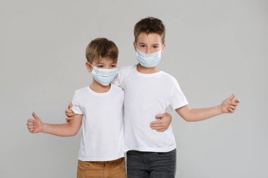 Beyaz elbiseli ve koruyucu maskeli iki sevimli çocuk parmak kaldırıyor, gri bir arka planda izole edilmiş..