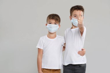 Beyaz elbiseli ve koruyucu maskeli iki sevimli çocuk parmak kaldırıyor, gri bir arka planda izole edilmiş..