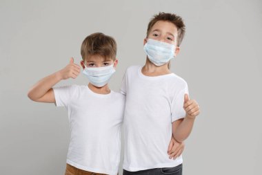 Beyaz elbiseli ve koruyucu maskeli iki sevimli çocuk parmak kaldırıyor, gri bir arka planda izole edilmiş..