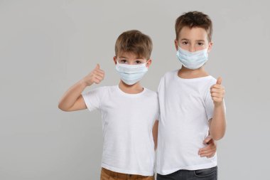 Beyaz elbiseli ve koruyucu maskeli iki sevimli çocuk parmak kaldırıyor, gri bir arka planda izole edilmiş..