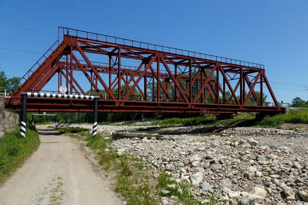 UTULIK, RUSIA - 15 JUNIO 2020: Puente ferroviario de metal de color rojo sobre un pequeño río de montaña con una carretera rural debajo — Foto de Stock