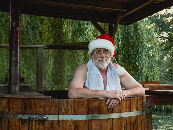 Senior man as Santa Claus with towel standing in a bath with warm water outside and looking with smiling at the camera.