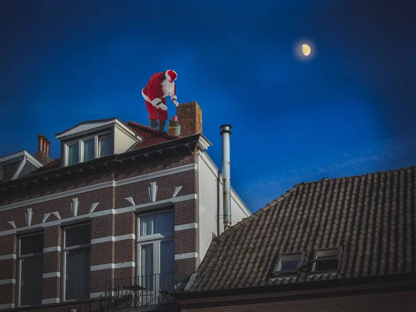 Santa Claus on the roof of a house puts the presents in the chimney. Christmas night scene, on the background of dark blue sky with a moon.