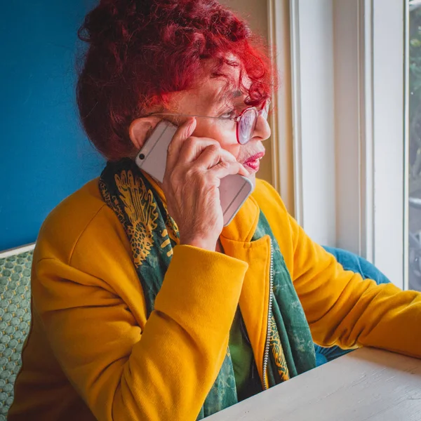 Modern older woman with red hair and glasses sitting before window holds phone in her hand and speaking.