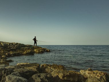 Genç bir adam bir taş kıyıdan denizde balık yakalar. Bir balıkçı ufukta silueti.