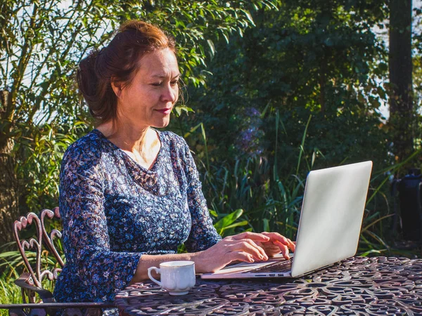 Femme souriante de 50 ans travaillant dans le jardin avec un ordinateur portable co — Photo