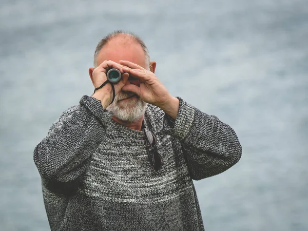 I see you. Senior man holding binoculars in his hands and looking at us. Using binoculars for better viewing.