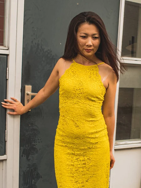 灰色の汚れたドアの前に立って お祝いの黄色のレースのドレスで長い髪を持つ中高年アジアの女性エレガントな — ストック写真