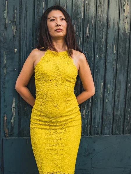 Pensive Asian model of 40 years in a yellow lace dress on the background of a rough textured wooden door.