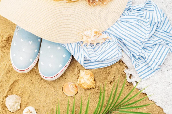 Top view of beach accessories on the sand. Hat, striped shirt, seashells, palm branches and slip-on sneakers on a tropical trip. Summer holidays.